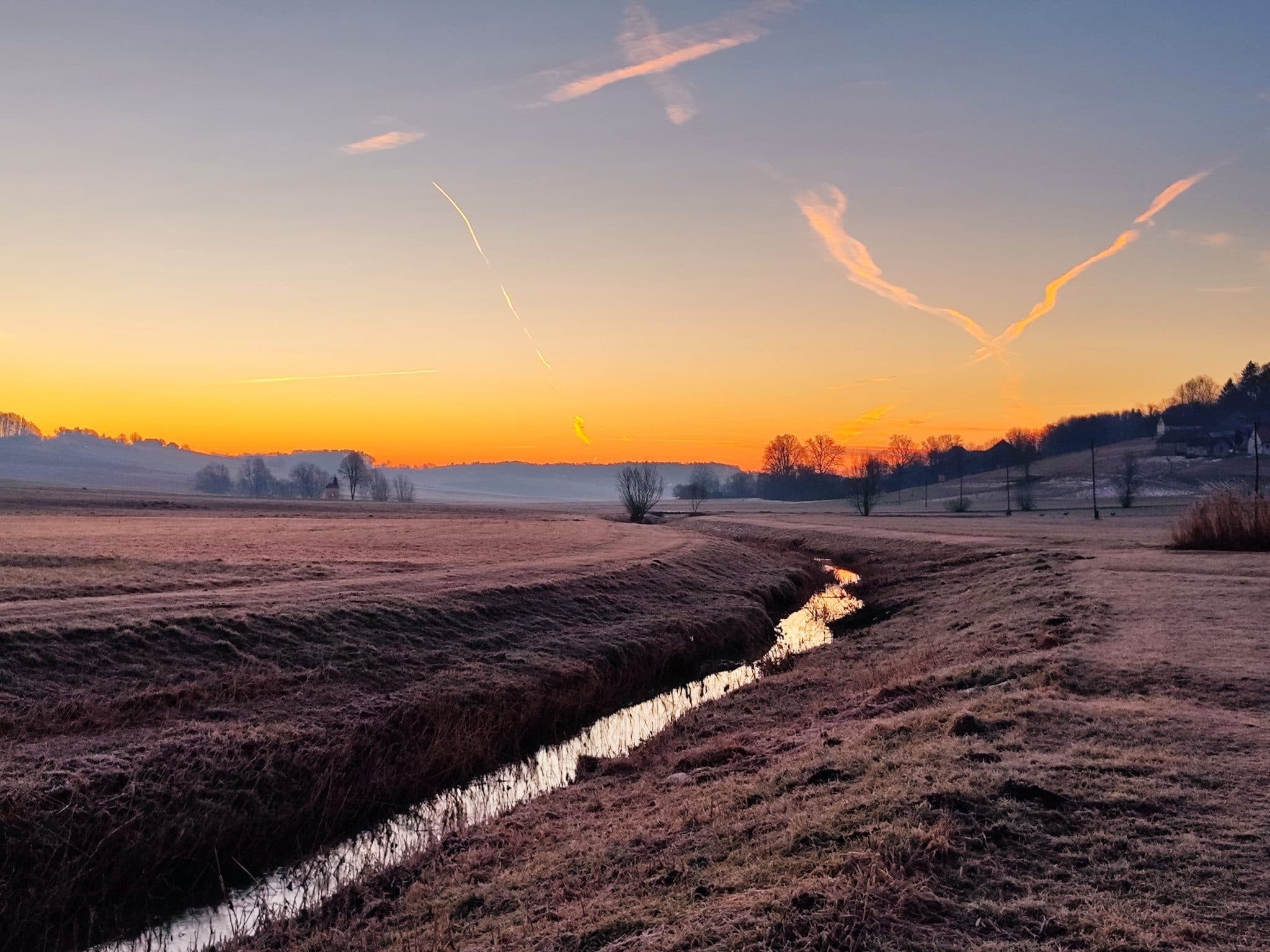 A narrow stream winding through a frosted meadow at first light, under an orange horizon.
